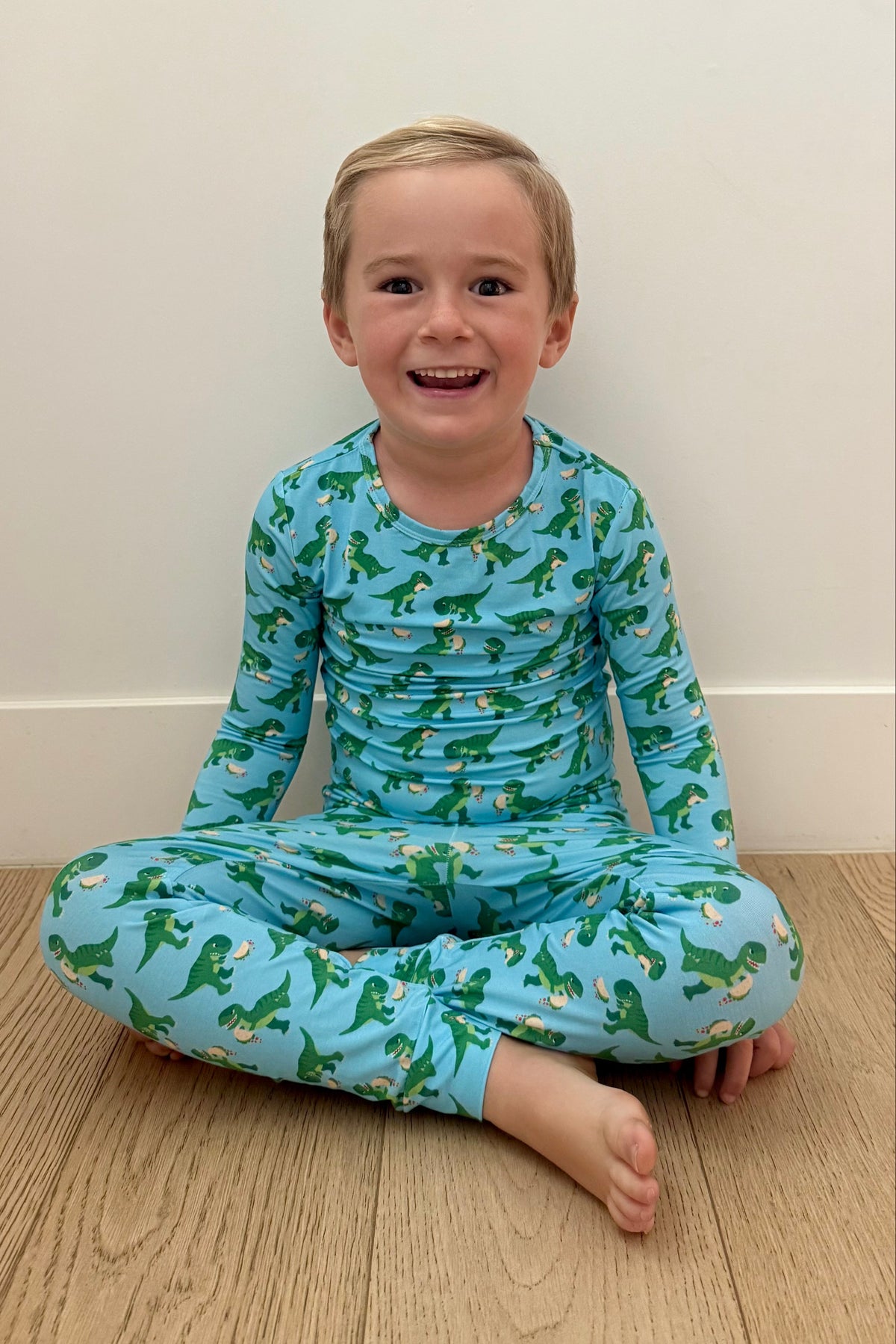 Child wearing blue pajamas with green dinosaur pattern sitting on a wooden floor.